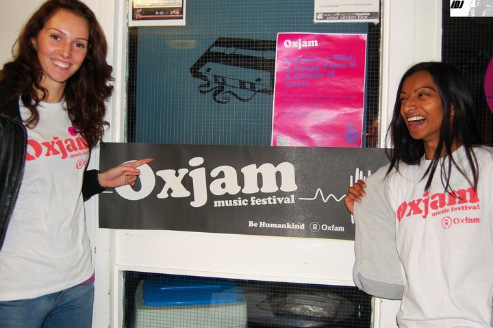 Two volunteers pointing at a black festival banner that says 'Oxjam', which is a local music festival event.